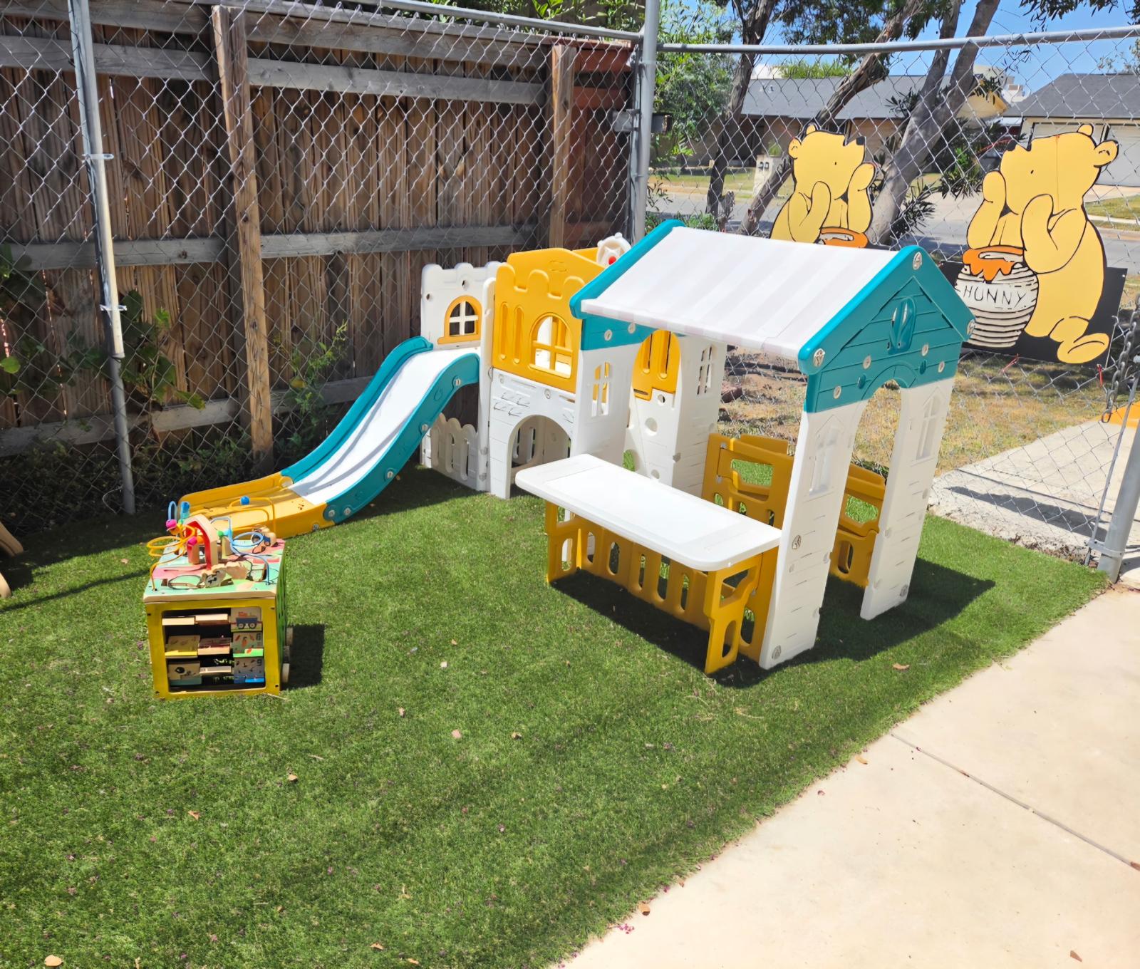 Turf play area with a yellow and teal playhouse, slide, bench, and wooden activity cube